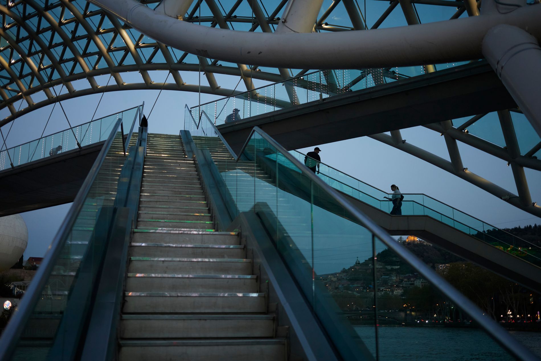 The Bridge of Peace illuminated at dusk — futuristic glass-and-steel pedestrian bridge over the Mtkvari River in Tbilisi