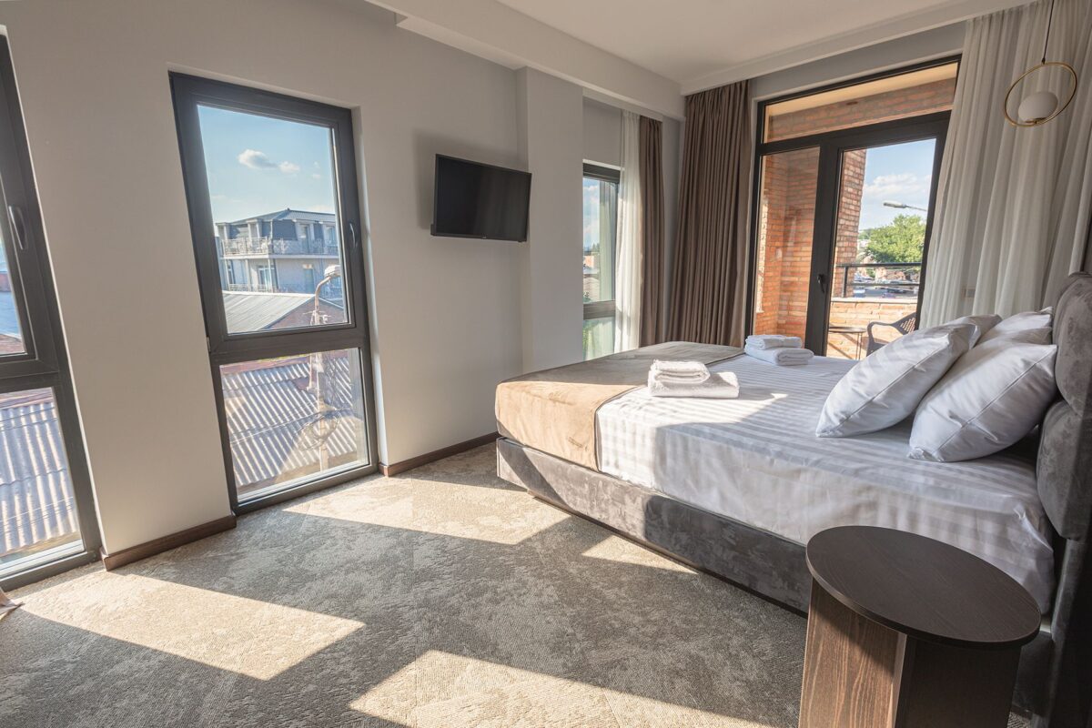 Sunlight entering through large balcony windows — cozy bed, TV and warm design in a premier cathedral-view room in Tbilisi